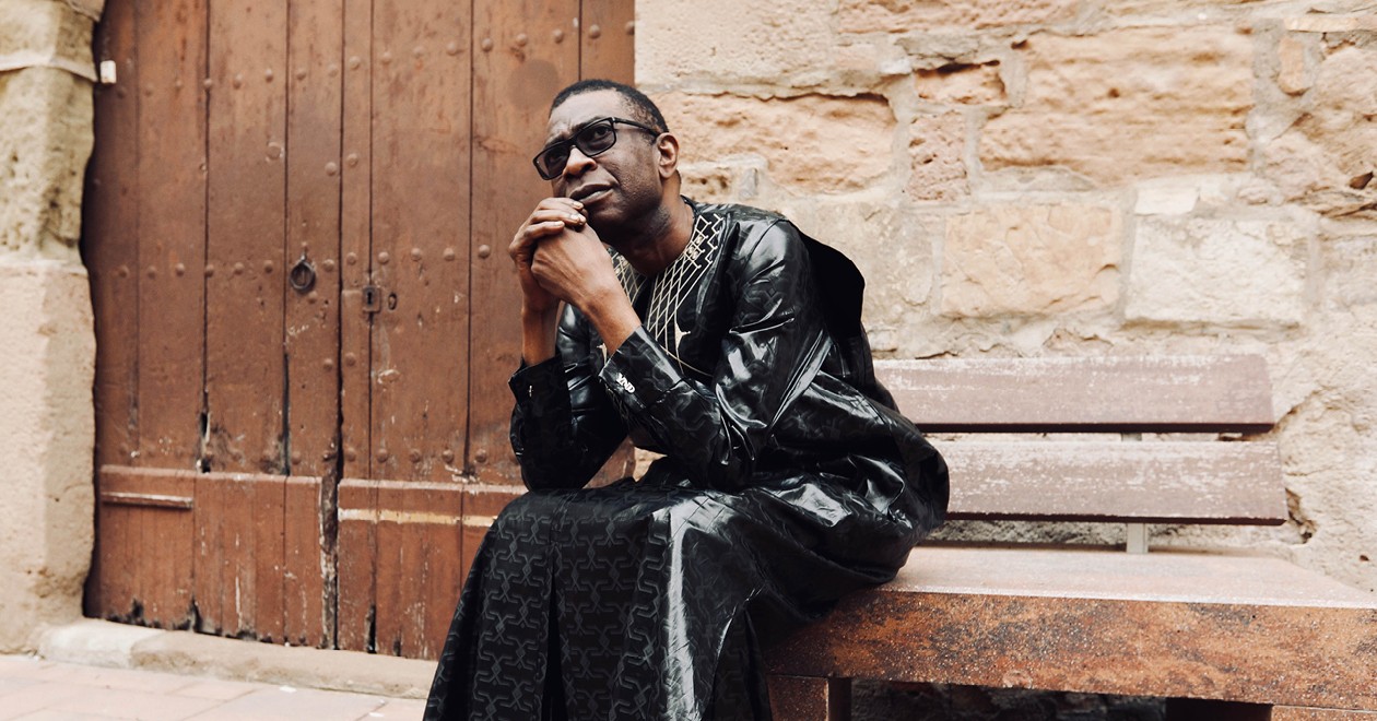 Youssou N'Dour seated on a bench in front of an old wooden door, wearing a black boubou